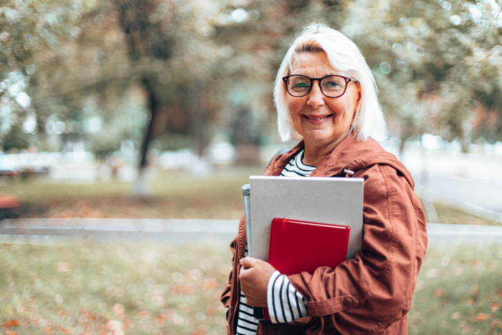 A senior woman holds books and a laptop on a college campus
